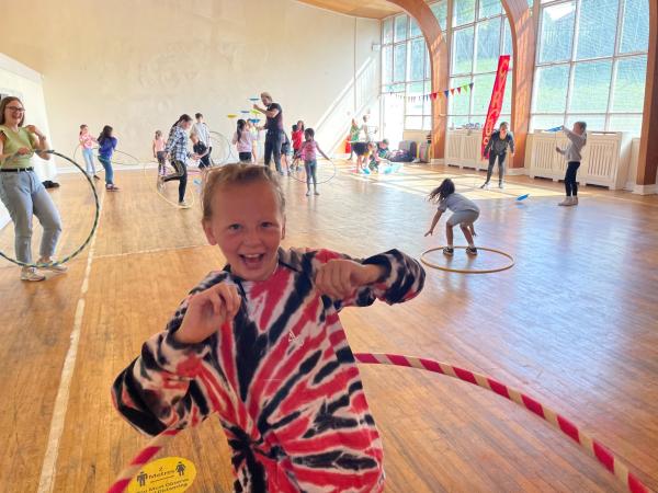 A group of kids are enjoying circus skills such as plate spinning and hula hoop, in a bright hall with bunting and flags in the backgroud