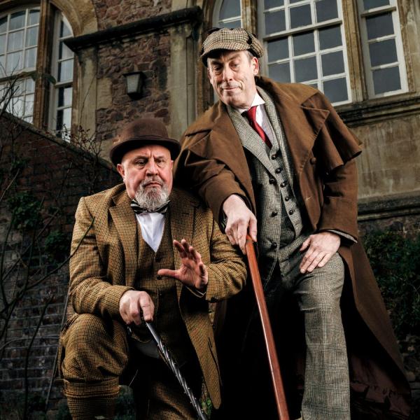 Peter Clifford and the Great Baldini portray a suited Holmes and Watson, who point their walking sticks towards a clue off-camera in the grounds of a grand, old-fashioned building.