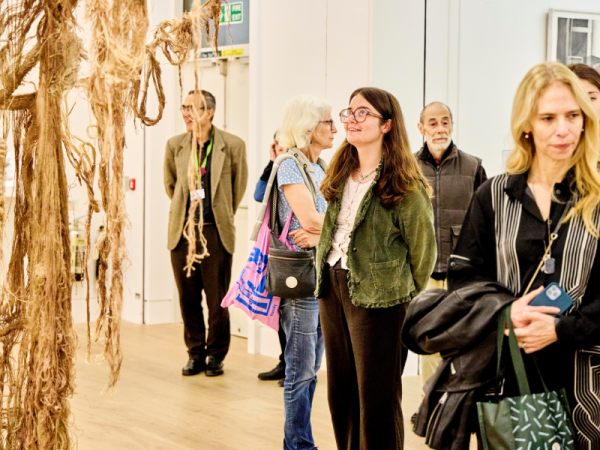 People gathered in a white walled gallery looking at the artworks. In the centre is a person with glasses wearing a green jacket looking up at a textile sculpture.