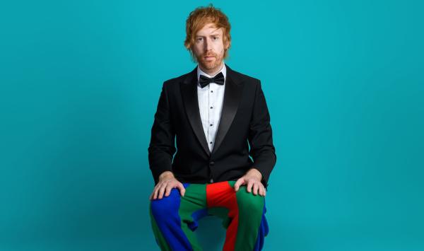 Ginger haired man sat wearing tuxedo jacket, bowtie and multi coloured jesters trousers. He could do with a shave