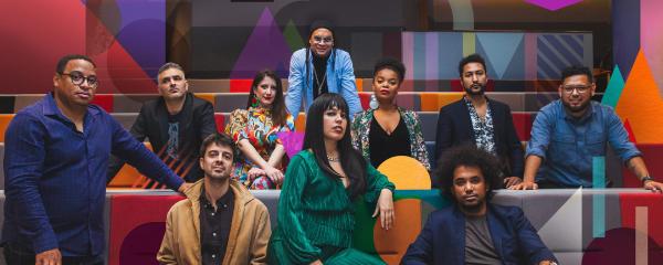 A group of ten people in stylish, artistic attire posing on tiered seating. The walls and furniture are decorated with vibrant, overlapping geometric patterns in a variety of colours.