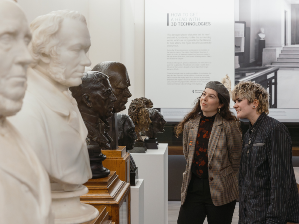 Two people looking at a row of portrait busts made of bronze and marble, stood on plinths