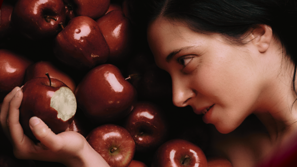 A person with long dark hair lying on some red apples. They are holding a bitten one in their hands.