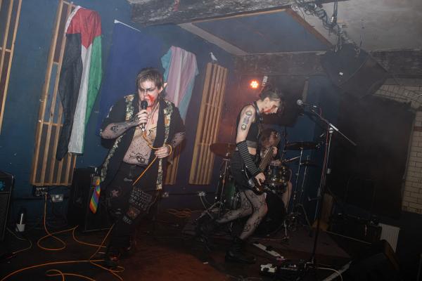 An image featuring two musicians on stage. In the background is a Palestine and Trans flag. 