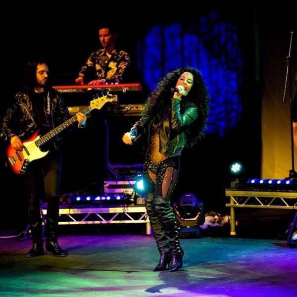 A Cher tribute performer sings into a handheld microphone beside a guitarist on stage. She has long, black, curly hair, leather jacket, knee-high boots and a glittery leotard.