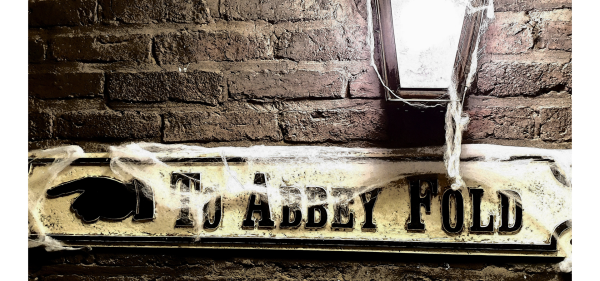 Image of a street sign saying ABBEY FOLD with black writing on a white background. A black finger points left. Brick wall behind it with a light on, and fake cobwebs giving a spooky feel.