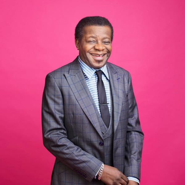 Wearing a grey suit in front of a pink background, Stephen K Amos stands laughing.