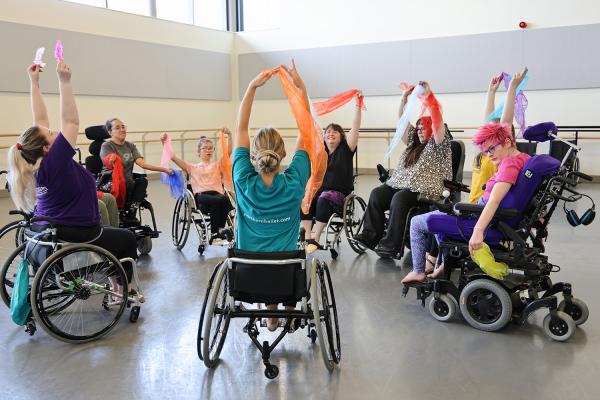 Wheelchair users in a big circle in a dance studio, raising theirs arms high with colourful scarves