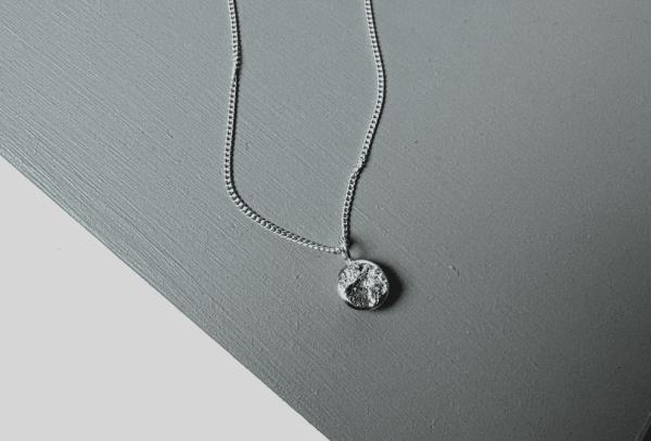 Image shows a textured ingot necklace on top of a grey tabletop. 