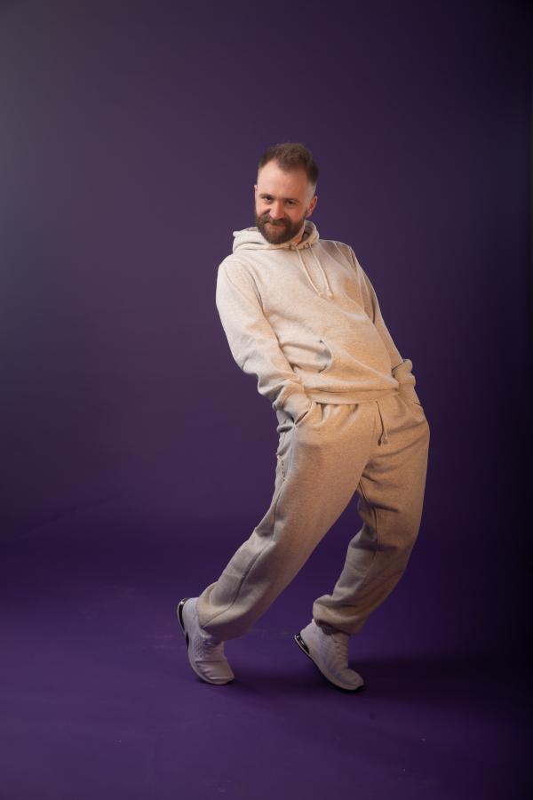 A white man with a grey tracksuit poses cheekily against a purple background.