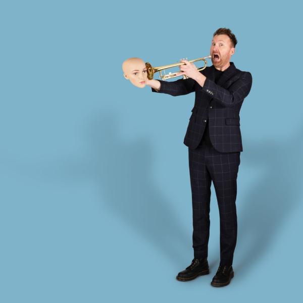 Jason Byrne in a suit comically plays a trombone, holding a mannequin's head to the instrument's bell.