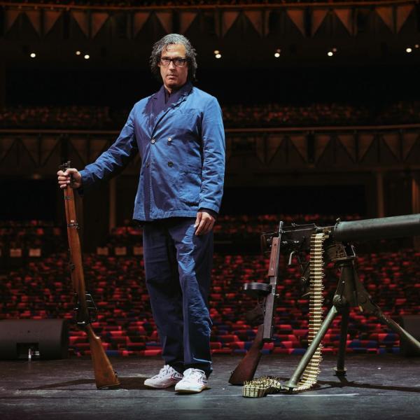 David Olusoga stands on stage in an auditorium, holding a vintage gun, beside two other old weapons.