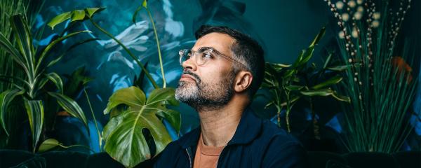 A man wearing glasses and a dark shirt stands in thought in front of a background of plants and vegetation.
