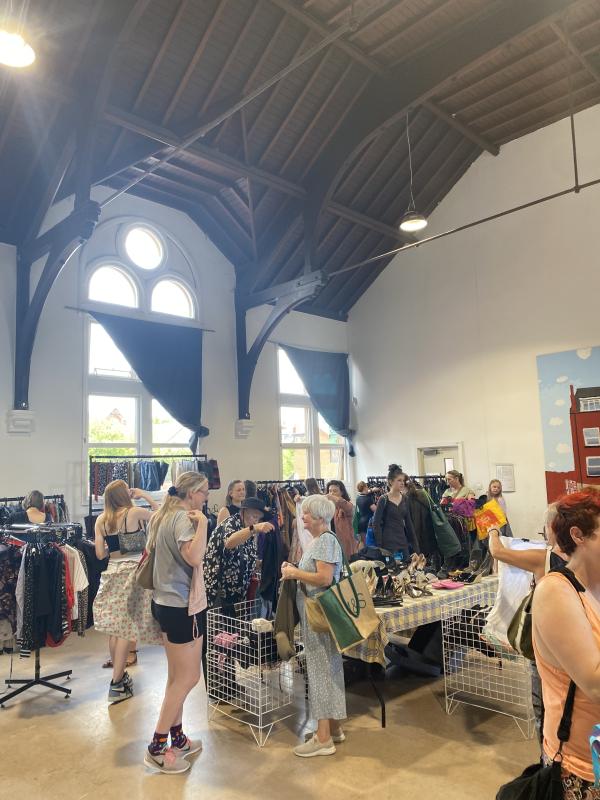 Groups of people browsing rails and holding up items of clothing in a brightly lit room filled with rails and baskets of clothing.