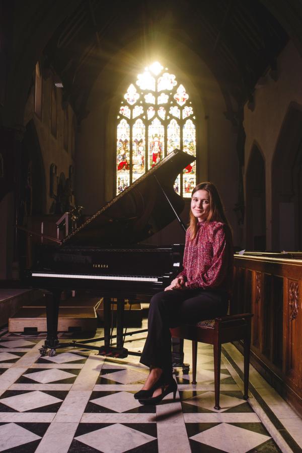 A lady sits on a chair in a church in front of a stained glass window there is a grand piano with its lid propped open the floor is a beautiful tiled pattern she's wearing high heels, I can't see where she's put her driving shoes