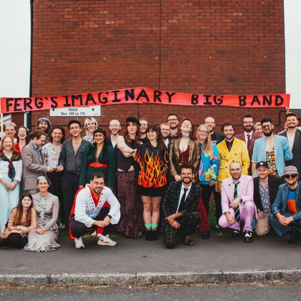 Ferg's Imaginary Big Band line up smiling on a street in front of a red brick building, adorned with a hand-painted banner that reads: Ferg's Imaginary Big Band.