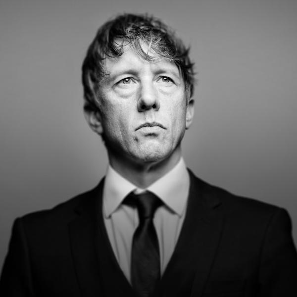 In black and white, Jonathan Pie looks sternly off-camera, standing poised in a black suit.