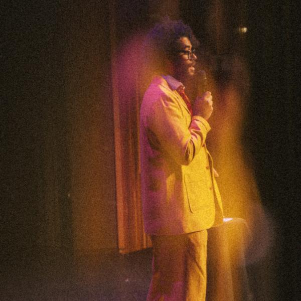 Grainy profile shot of Richard Ayoade stood on stage in a suit and tie, speaking into a microphone.