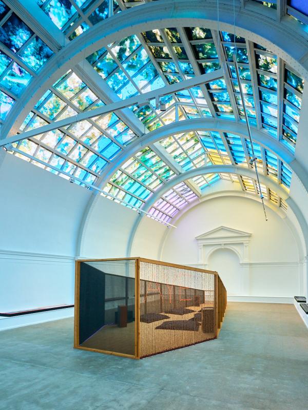 A large white room with a colourful lit ceiling above a wooden coffin shaped oversized sculpture.