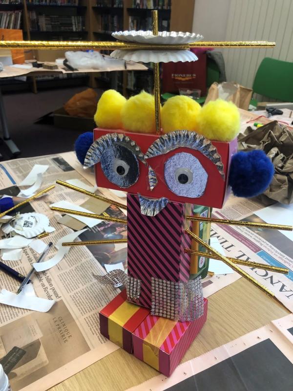 A small 'robot' made from junk materials with yellow pom pom hair, gold straw arms, shiny tin foil eyes and eye brows and stripey body.