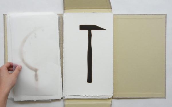 An open portfolio containing several prints, with a hand leafing through them. The top-most print is of a hammer, which has been burnt into the thick watercolour paper using a heated steel stencil.
