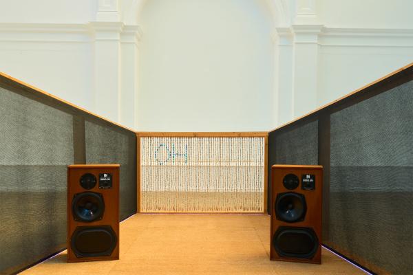  symmetrical gallery installation featuring two large vintage-style wooden speakers facing down a narrow corridor lined with tall dark acoustic panels. At the far end, a woven screen partially reveals the blue letters "OH." The installation sits within a bright white room with high ceilings, classical columns, and an arched alcove, creating a minimalist, immersive listening environment.