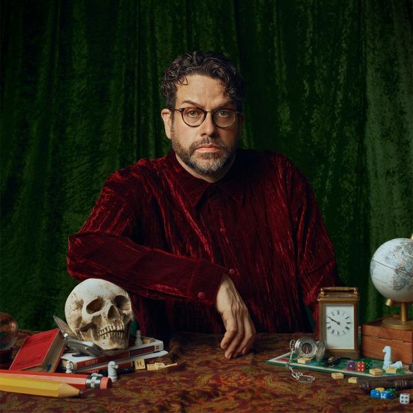 In the style of a Renaissance painting, Michael Spicer sits at a desk with his arm resting on a skull, as he wears a velvety red outfit in front of a dark green curtain. The desk also features pencils, books, dice, keys, a stopwatch and a globe.