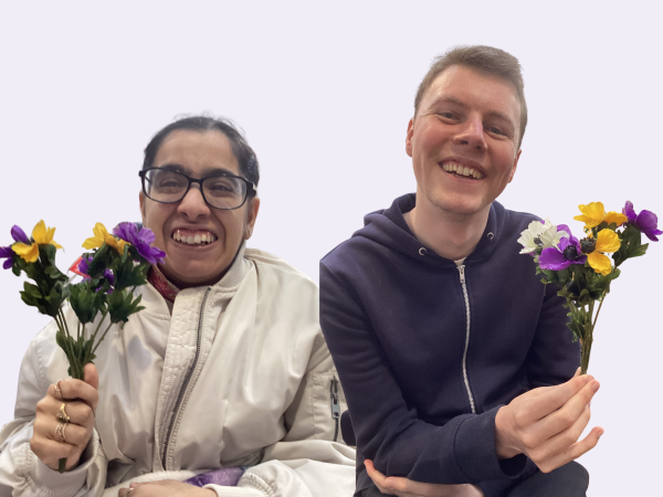 A woman and man are smiling at the camera and each holding a small bunch of yellow, purple and white flowers. They are against a lilac background.