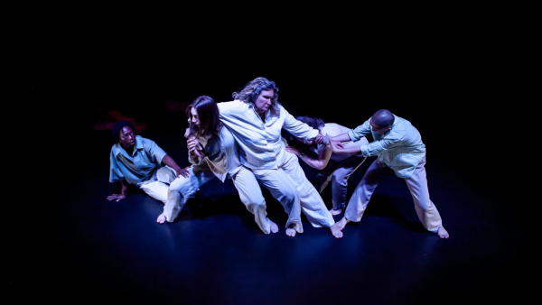 Four dancers perform in darkness, all crouching down. The dancer in the middle leans on the others. One dancer is sat, watching from behind them. 