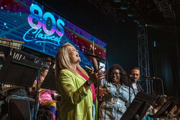 A group of singers performs energetically on stage in front of an orchestra, with a large "80s Classical" sign in the background.