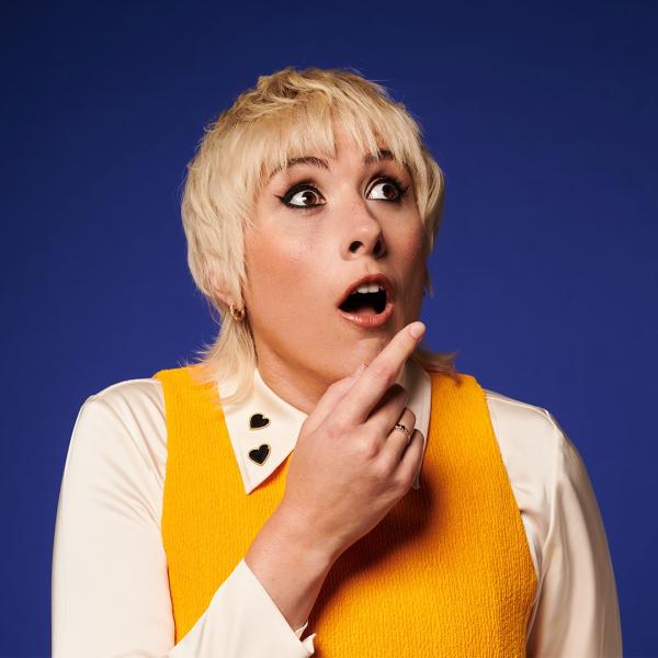 With a shocked expression and bleached blonde hair in a wide-collared shirt, Maisie Adam looks wide-eyed and open-mouthed as she points off-camera.