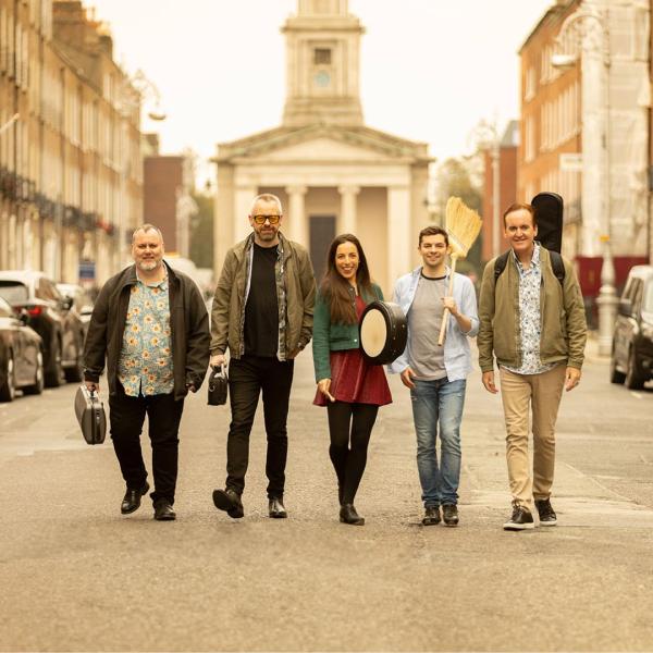 On a sunny street, the members of Irish House Party smile and walk towards the camera, each carrying an instrument case, one holding a broom.