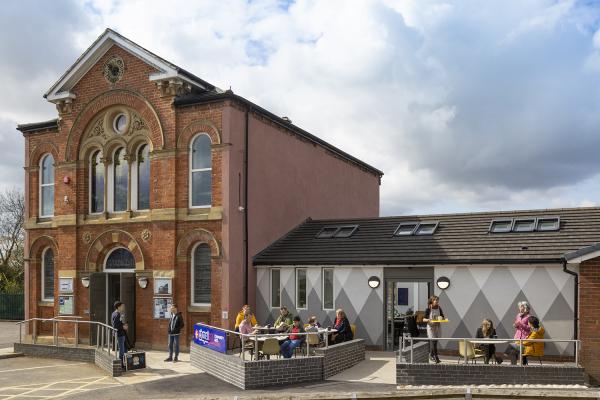 A red-brick Victorian chapel, now the Chapel FM Arts Centre, features ornate arched windows and a decorative gable. To the right, a modern one-story extension with a distinctive grey-and-white diamond pattern on its exterior wall includes a small paved terrace where people are seated at outdoor tables. A staff member is seen carrying a tray toward the patrons under a bright, cloudy sky.