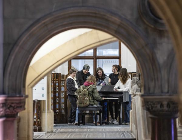 A view through a stone arch shows a group of young people around a piano.