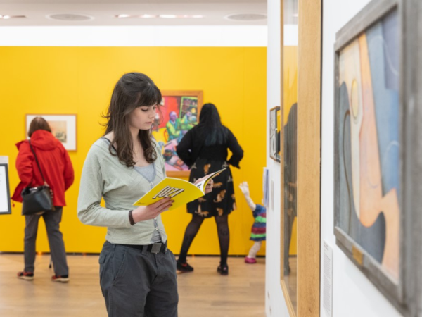 A photo of someone with long black hair wearing a pale green cardigan looking at a yellow catalogue whilst stood in an exhibition. The wall behind is painted yellow.