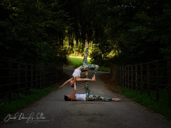 Two women in a park, wshrouded by the shade of overhead trees on a path, they are posed one holding the other up in the air. An opening to light through the trees is in the background