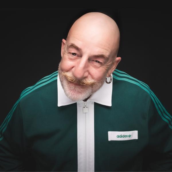Jeff Innocent, a bald man with a beard and moustache, leans towards the camera, smirking. He wears a dark green Adidas tracksuit with a white collar.