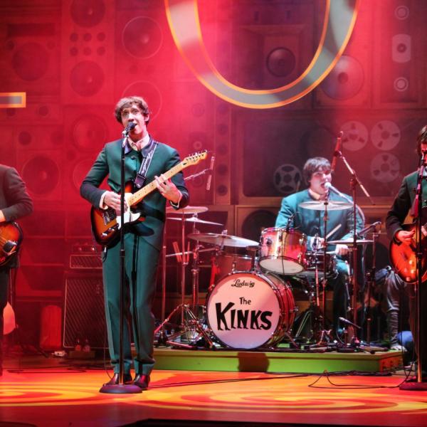 Production shot from Sunny Afternoon of someone playing an electric guitar and drummer with The Kinks logo on the bass drum.