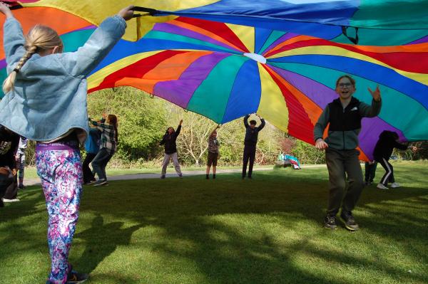 A group of children playing a parachute game with a rainbow striped parachute.