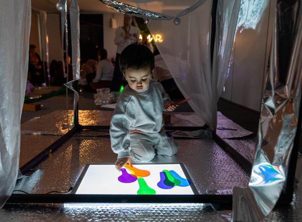 A child in a grey outfit interacts with a brightly lit color panel, touching colorful shapes in a dimly lit, reflective room filled with people.