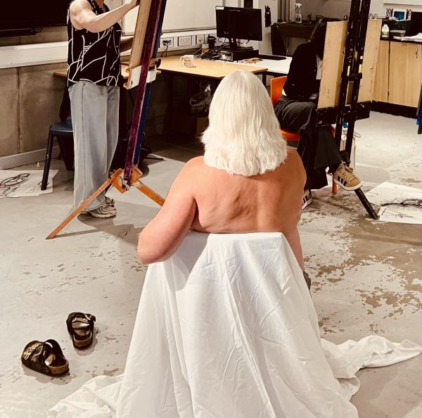 A life model sits with her back to the camera and in front of her a student sketches