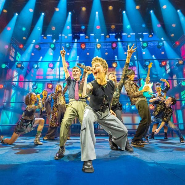 A band of musicians play on stage under bright lighting, led by a person singing into a microphone. The stage is full with multicoloured spotlights.