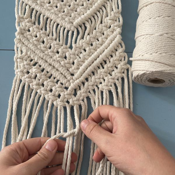 A photograph showing a pair of hands tying a knot on a WIP of a beige macrame wall hanging
