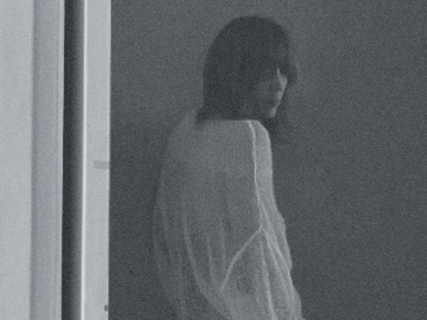 A grainy, black and white image of a woman with long dark hair, wearing a long white shirt.