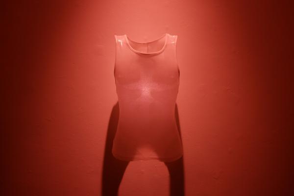 A translucent sleeveless vest hangs against a deep red wall illuminated by red light. 