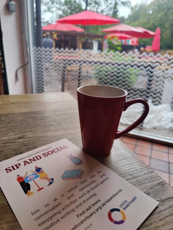 Image shows a red mug of tea on a table, next to a flyer for Sip and Social, near a window. A courtyard with benches and red umbrellas can be seen outside.