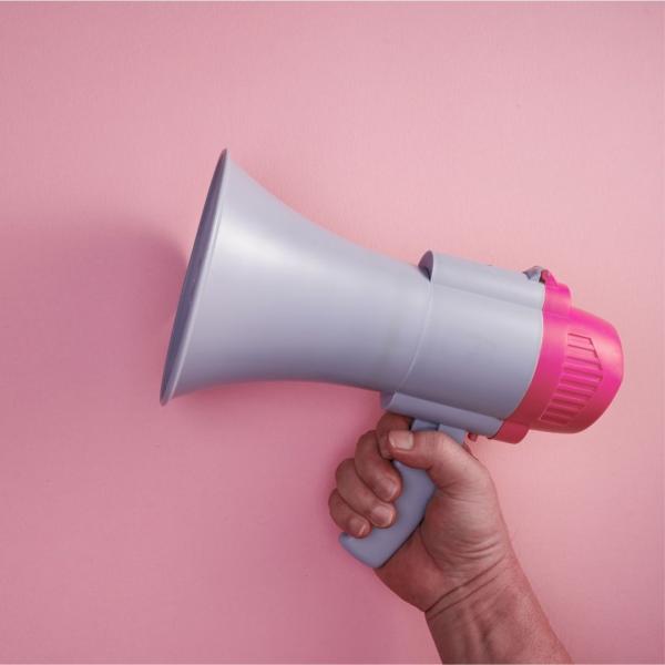 A photograph of a hand holding a white megaphone against a pink background
