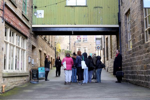 A group of visitors on a Heritage Tour of Sunny Bank Mills. They are gathered on the Mills' main hill under a green painted wooden clad bridge, connecting two Yorkshire stone built mill buildings.