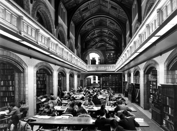 Black and white photograph of a library. Long tables are filled with people studying.