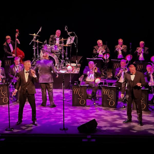 A big band on stage, featuring a drummer, cellist and many men with trumpets, saxophones and trombones perform, led by two men singing in tuxedos.